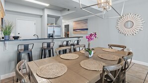 A dining area with a long table, wooden chairs, woven placemats, a vase with flowers, and a modern chandelier. - Beach Front Unit - Tops'l Resort - Free Pickleball - Tides 1304 (Miramar Beach)