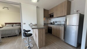 Una cocina moderna con armarios de madera, un refrigerador de acero inoxidable y un microondas. Hay una pequeña isla con taburetes altos y una cama en la habitación contigua.