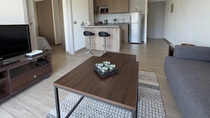 Una sala de estar moderna con televisor, una mesa de centro y un sofá. Se aprecia una cocina al fondo con refrigerador, microondas y taburetes de bar.