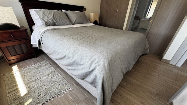 Een net opgemaakt bed met een grijze deken, een houten nachtkastje met een lamp en een geweven vloerkleed op een houten vloer.