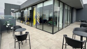 Een modern gebouw met grote glazen deuren, een buitenzitplaats met tafels en stoelen, en een stadsgezicht op de achtergrond.