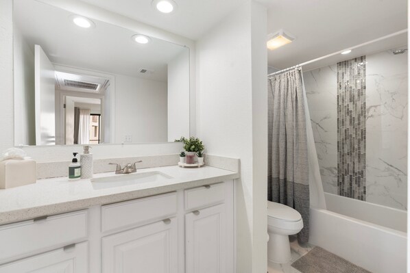 Un baño moderno con un tocador blanco, un espejo grande, un inodoro y una zona de ducha con pared de mármol y cortina.