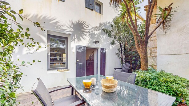 An outdoor dining area with a glass table, chairs, and a small garden with plants and a tree.