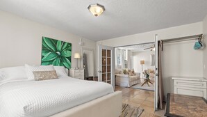 A bedroom with a large bed, a green abstract painting, a nightstand, a dresser, and a view into another room with a sofa and a lamp. - Ocean View Mid Waikiki 1 Block to the Beach (Honolulu)
