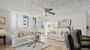 A modern living room with two white sofas, a glass coffee table, a ceiling fan, and a view of a building outside. - Ocean View Mid Waikiki 1 Block to the Beach (Honolulu)