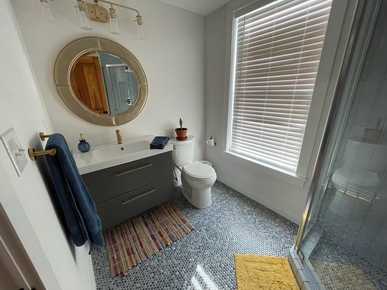 Ein Badezimmer mit rundem Spiegel, Toilette, Waschbecken und Handtuchhalter sowie einem Fenster mit Jalousien.