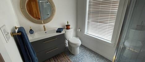 Ein Badezimmer mit rundem Spiegel, Toilette, Waschbecken und Handtuchhalter sowie einem Fenster mit Jalousien.