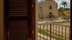 Une vue depuis un balcon avec une porte en bois, donnant sur un bâtiment historique doté d’une tour et de flèches, une rue bordée de palmiers et un ciel dégagé.