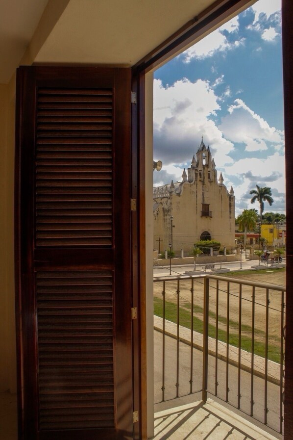 A view from a balcony with a wooden door, overlooking a historic building with a tower and spires, a street with palm trees, and a clear sky. - Casona de la Guayabera Hospedaje  (Tekit)