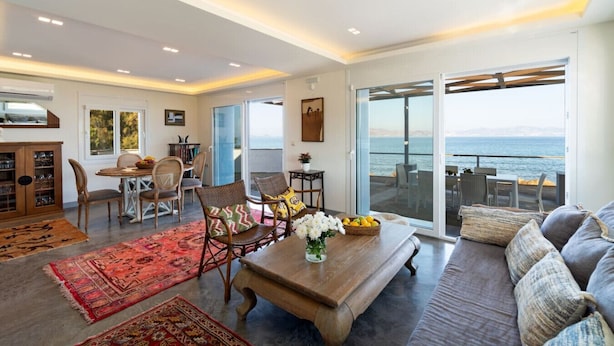 Un vaste espace de vie avec une grande fenêtre donnant sur la mer, un coin repas avec des meubles en osier et un coin salon confortable avec un canapé et des coussins.