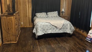 A bedroom with a wooden floor, a bed with patterned bedding, a dresser, a television on the dresser, and a bag of chips on the floor. - Cozy 2-bedroom cabin with WiFi, AC in charming Murfreesboro on Lake Greeson. (Murfreesboro)
