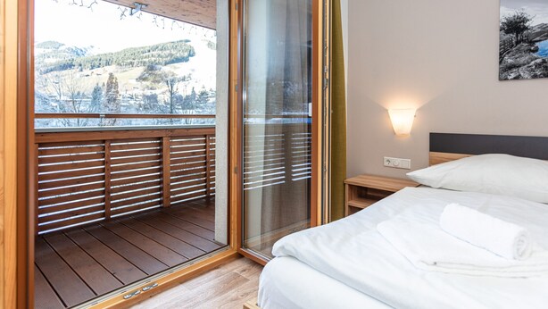 Un dormitorio con un amplio ventanal corredera que da a un balcón, una cama con ropa blanca, una mesita de noche y una lámpara de pared.