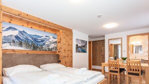 Una cama de madera con dos almohadas y un edredón ordenado, un escritorio de madera con una silla, y un cuadro grande de un paisaje montañoso nevado en la pared.