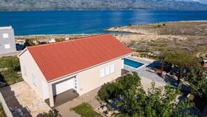 Een modern huis met een rood dak, een zwembad en uitzicht op een groot wateroppervlak en bergen op de achtergrond.