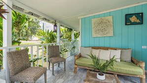 Une véranda couverte, agrémentée de fauteuils en osier et d’un banc en bois, entourée d’une végétation tropicale luxuriante.