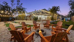 A patio area with wooden Adirondack chairs arranged around a fire pit, a glass greenhouse structure, and a small putting green.