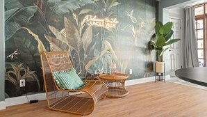 A room with a mural of a jungle, a wicker chair with a striped pillow, a round wooden table, and a large potted plant.
