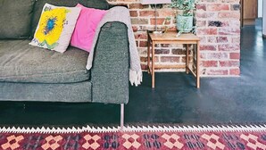A room with a brick wall, a sofa with cushions, a small wooden side table with a lamp and a plant, and a hanging light fixture.