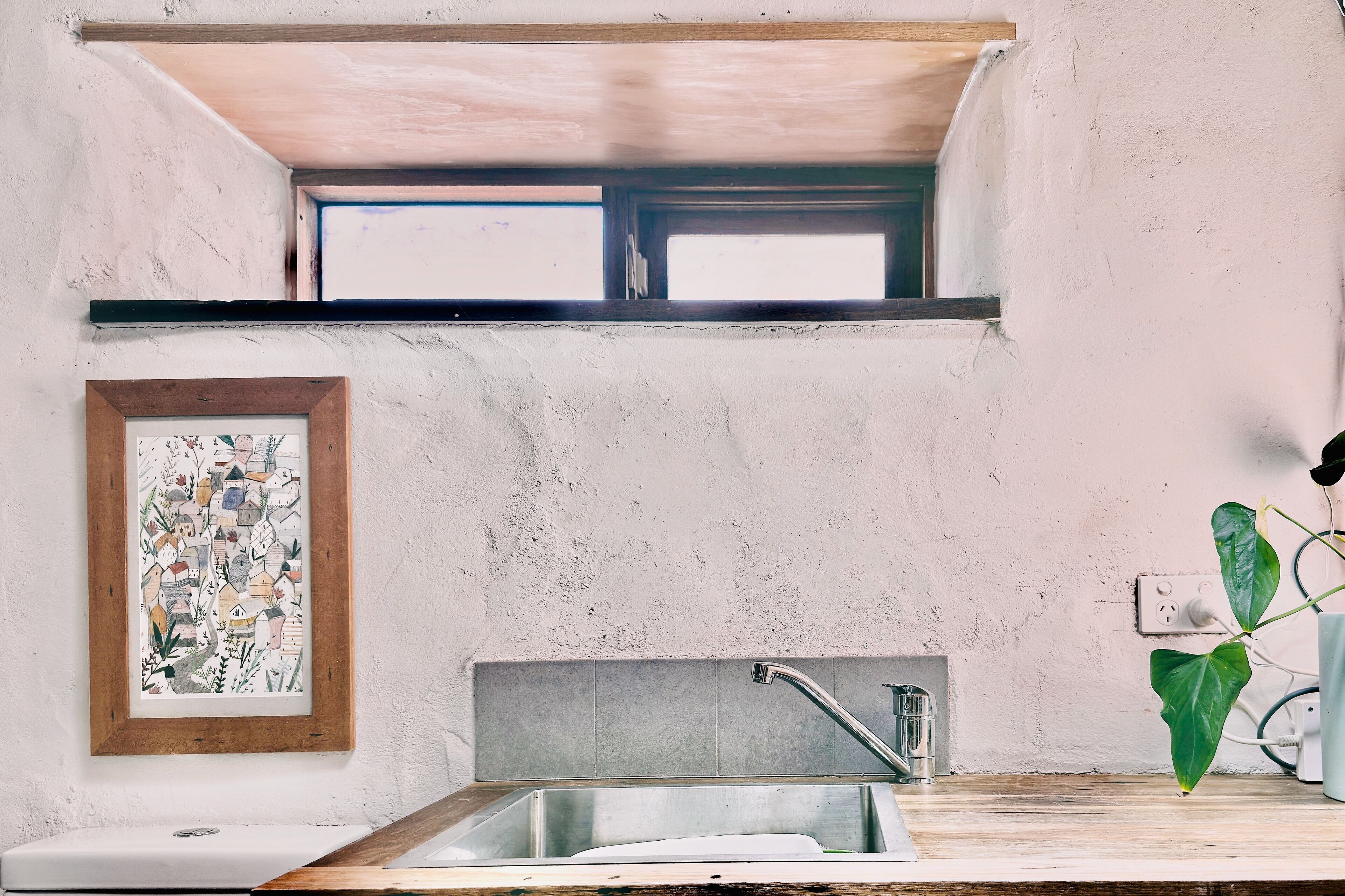 Una cocina con ventana, un lavamanos, un cuadro enmarcado y una planta.