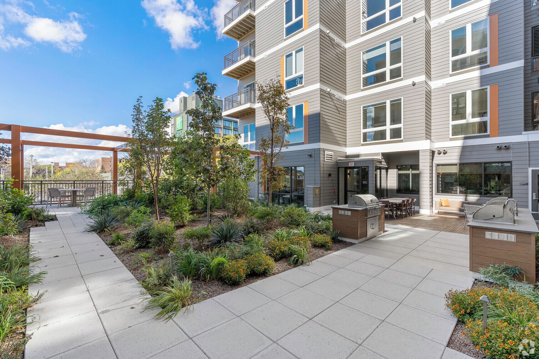 A modern apartment building with a patio area featuring outdoor seating, a grill, and a landscaped garden.