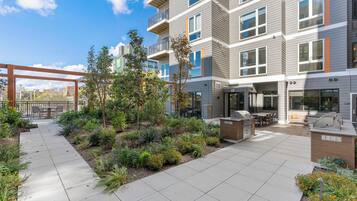 A modern apartment building with a patio area featuring outdoor seating, a grill, and a landscaped garden.