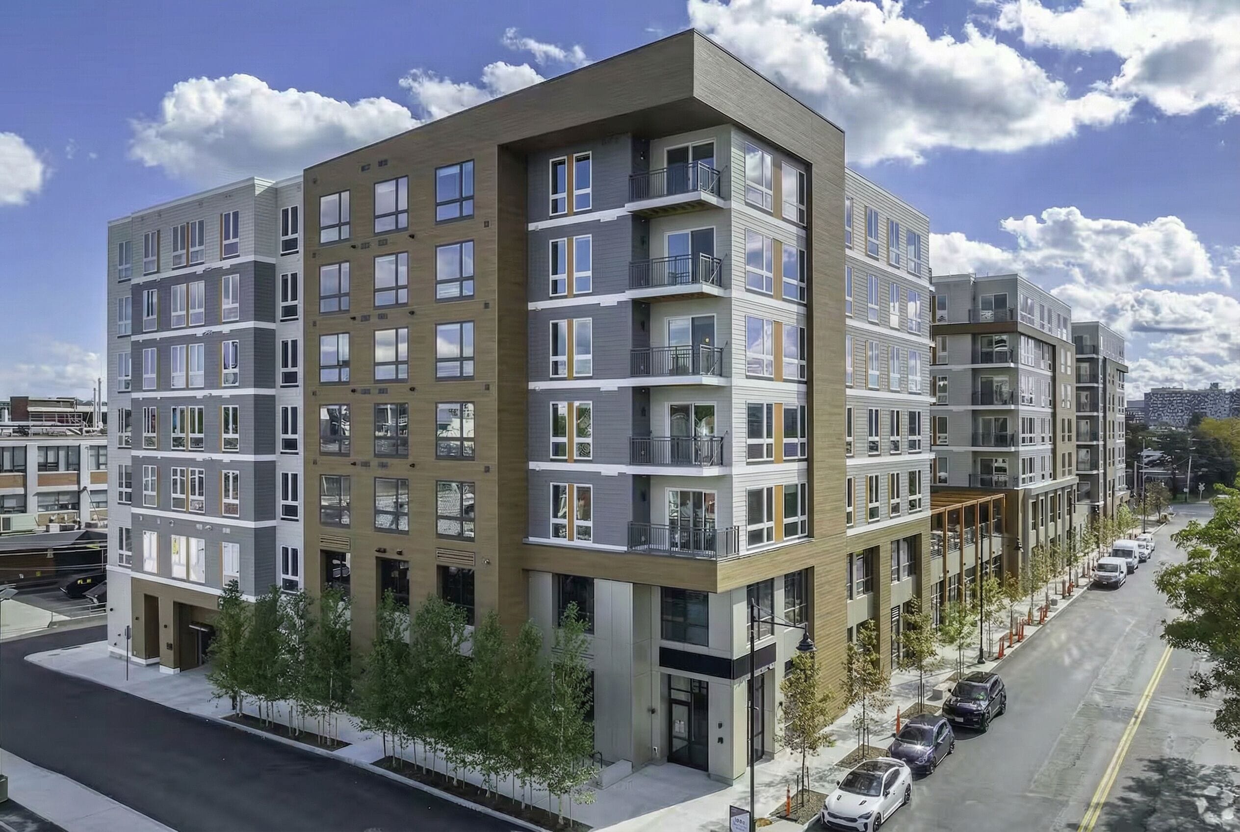 A multi-story residential building with balconies, a parking area, and a clear sky.