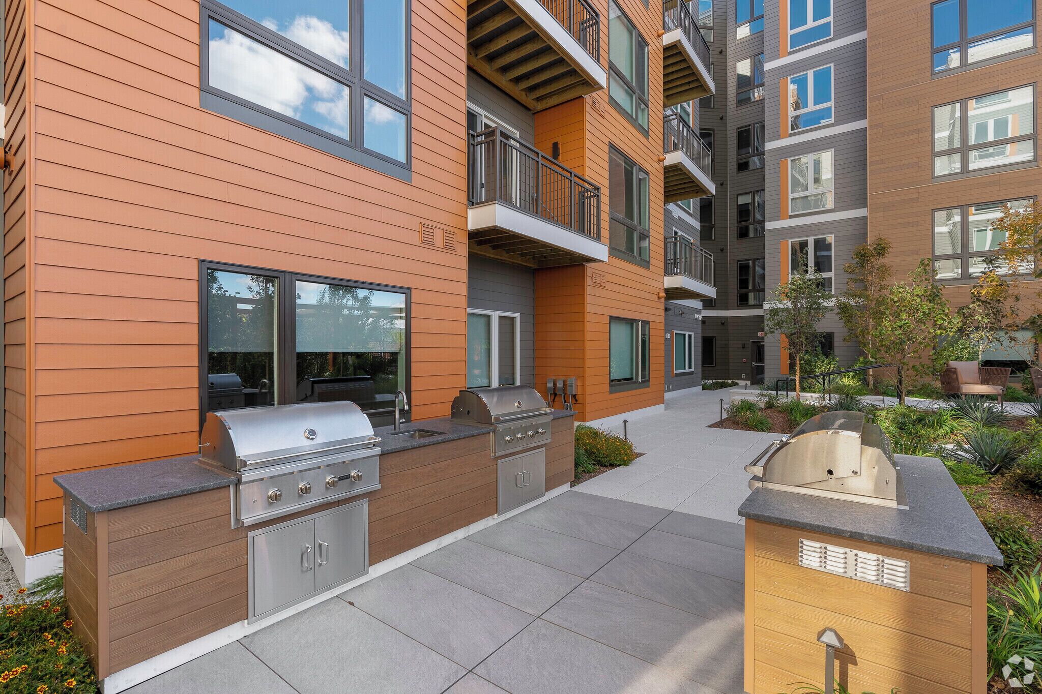 Un área de cocina exterior moderna con una parrilla, armarios empotrados y una estación de barbacoa, junto a un edificio residencial de varios pisos con balcones.