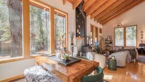 Amplia sala de estar con una mesa de comedor de madera, asientos cómodos, una chimenea y ventanas grandes que ofrecen vistas del bosque circundante.
