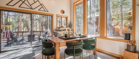 A spacious dining area with a wooden table and green upholstered chairs, large windows offering a view of the forest, and a modern ceiling light fixture.