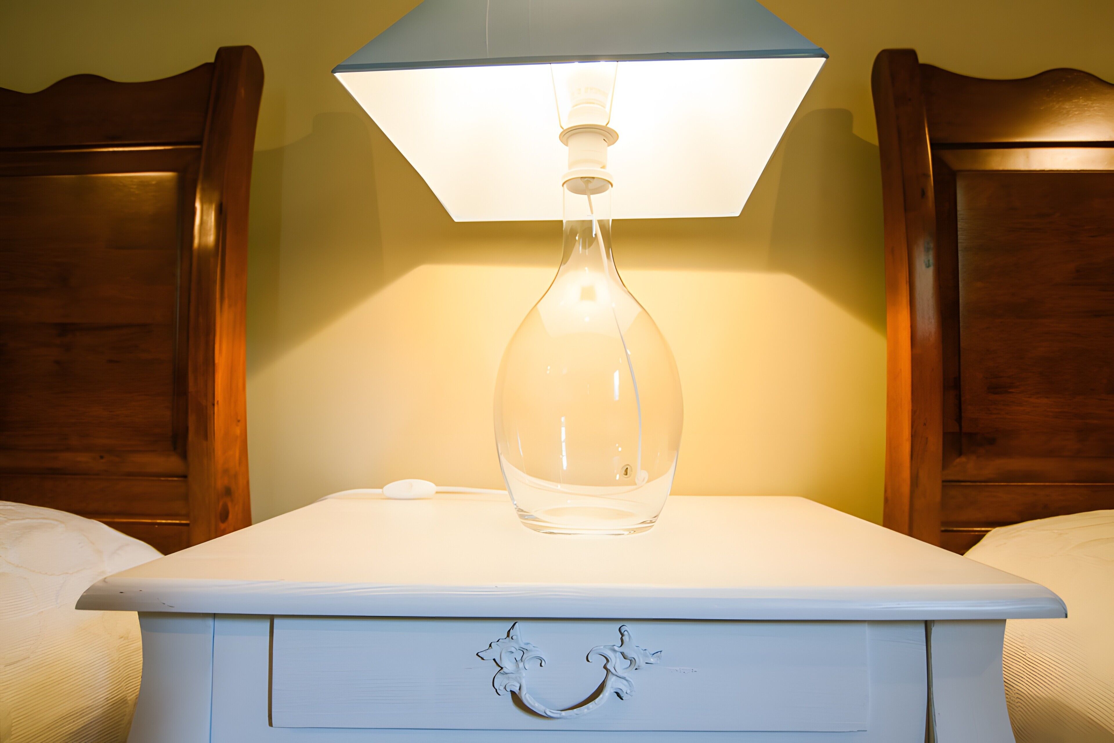 A white bedside table with a clear glass bottle and a lamp on top, wooden headboard, and a drawer with a handle.