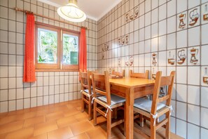 Een eetgedeelte met een houten tafel en stoelen, een raam met gordijnen en een betegelde muur met illustraties van keukengerei.