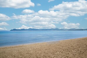 Een strand met helder water, verre bergen en een breed zandstrand.