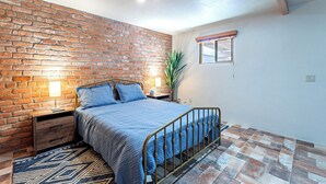 Een slaapkamer met een bakstenen muur, een metalen bed, een blauw beddegoed, een nachtkastje met een lamp en een raam met uitzicht op buiten.