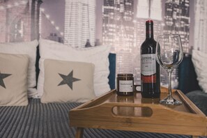 Una bandeja de madera con una botella de vino, una copa y una vela sobre una cama con almohadas estampadas con estrellas y vistas a la ciudad.