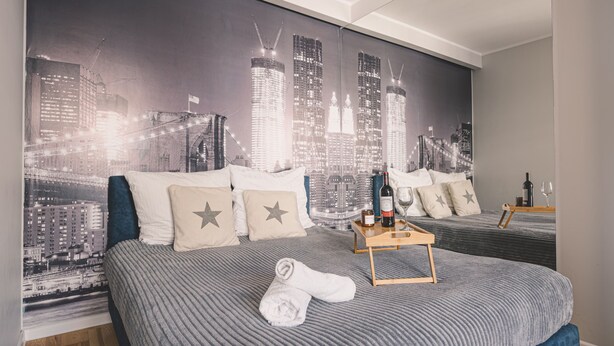 A hotel room with a cityscape mural on the wall, a bed with a grey striped bedspread, two pillows, a wooden nightstand with a bottle and glasses, and a rolled towel on the bed. - Dream 18 Przytulne Studio Centrum Bydgoszczy (Bydgoszcz)