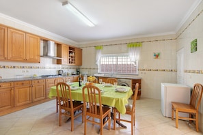A kitchen with wooden cabinets, a dining table with chairs, a refrigerator, and a window with curtains.