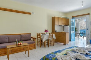 Een kamer met een bank, een eettafel met stoelen, een keukenhoek en uitzicht door een open deur.