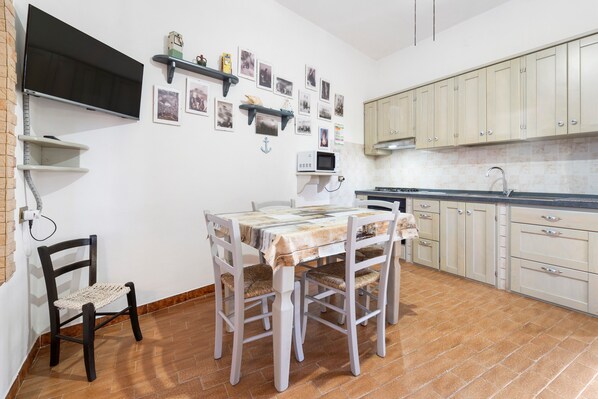 Una cocina con armarios blancos, suelo de baldosas, una mesa de comedor con sillas, un televisor montado en la pared y diversas fotos y adornos.