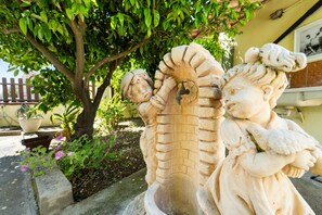 Une sculpture en pierre représentant une figure humaine avec un serpent, placée dans un jardin agrémenté d’un arbre et de plantes en pot.