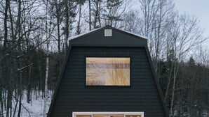 A dark-colored cabin with a wooden deck, large windows, and a visible interior with a fireplace.