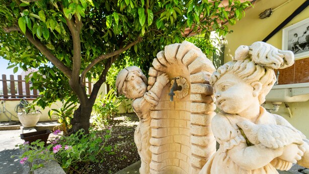 Escultura de piedra de una figura con una serpiente, ubicada en un jardín con un árbol y plantas en macetas.