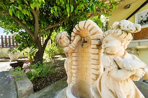 Eine Steinskulptur einer Figur mit einer Schlange, eingebettet in einen Garten mit einem Baum und Kübelpflanzen.