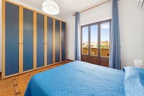 Ein Schlafzimmer mit Bett, Schrank, Fenster mit Vorhängen und Blick auf Gebäude.