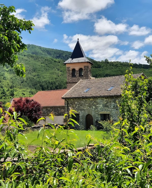 A stone building with a red roof and a bell tower, surrounded by lush greenery and a clear sky.