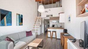 A compact living space with a sofa, a small table, a kitchen area with a counter and stools, a staircase, and a TV. - AS25806-P2 duplex cabin with terrace and parking (LE GRAU DU ROI)