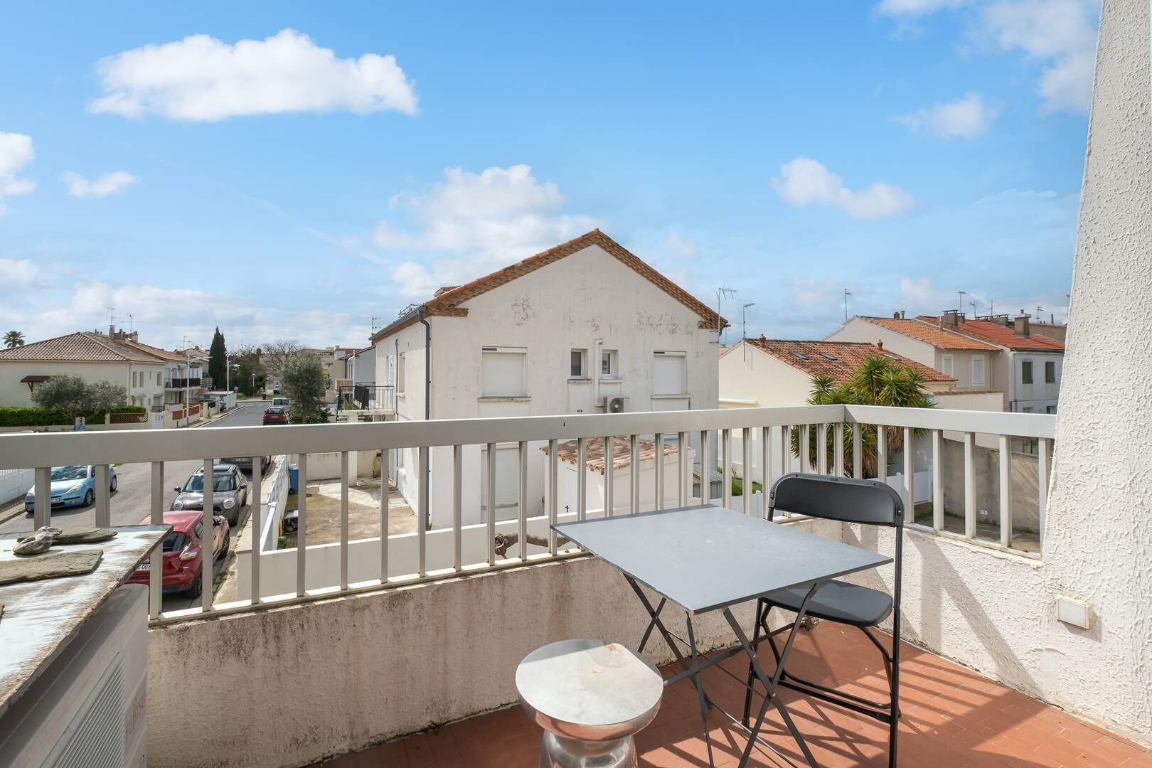 A balcony with a table and chair, overlooking a street with parked cars and buildings.