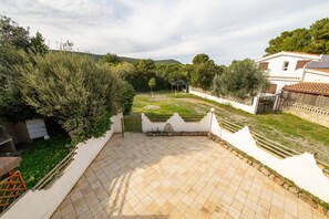 Une cour avec un sol carrelé, un mur blanc et un jardin agrémenté d’arbres et d’arbustes.