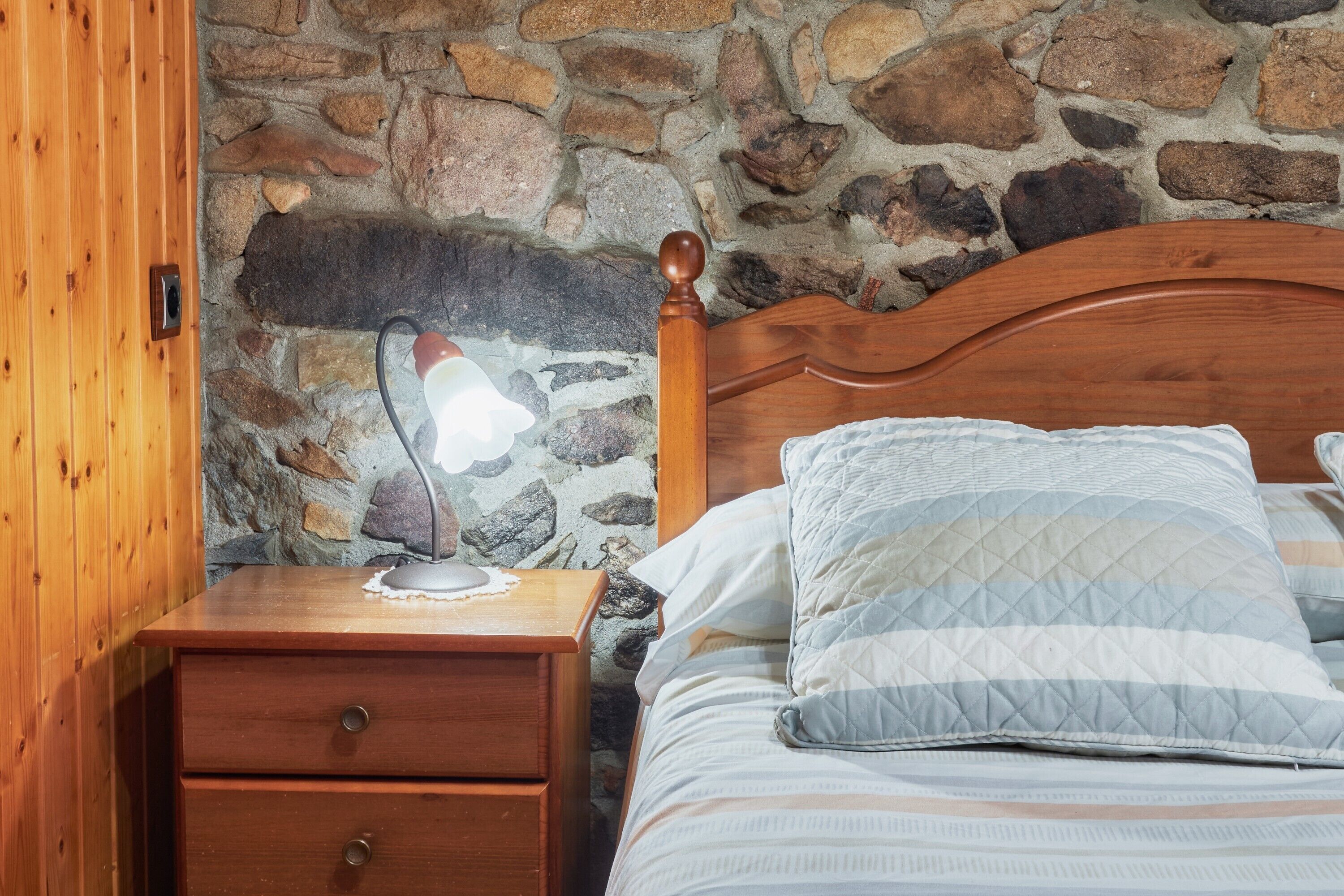 Una mesita de noche de madera con una lámpara, una cama con ropa de cama a rayas y una pared de piedra.