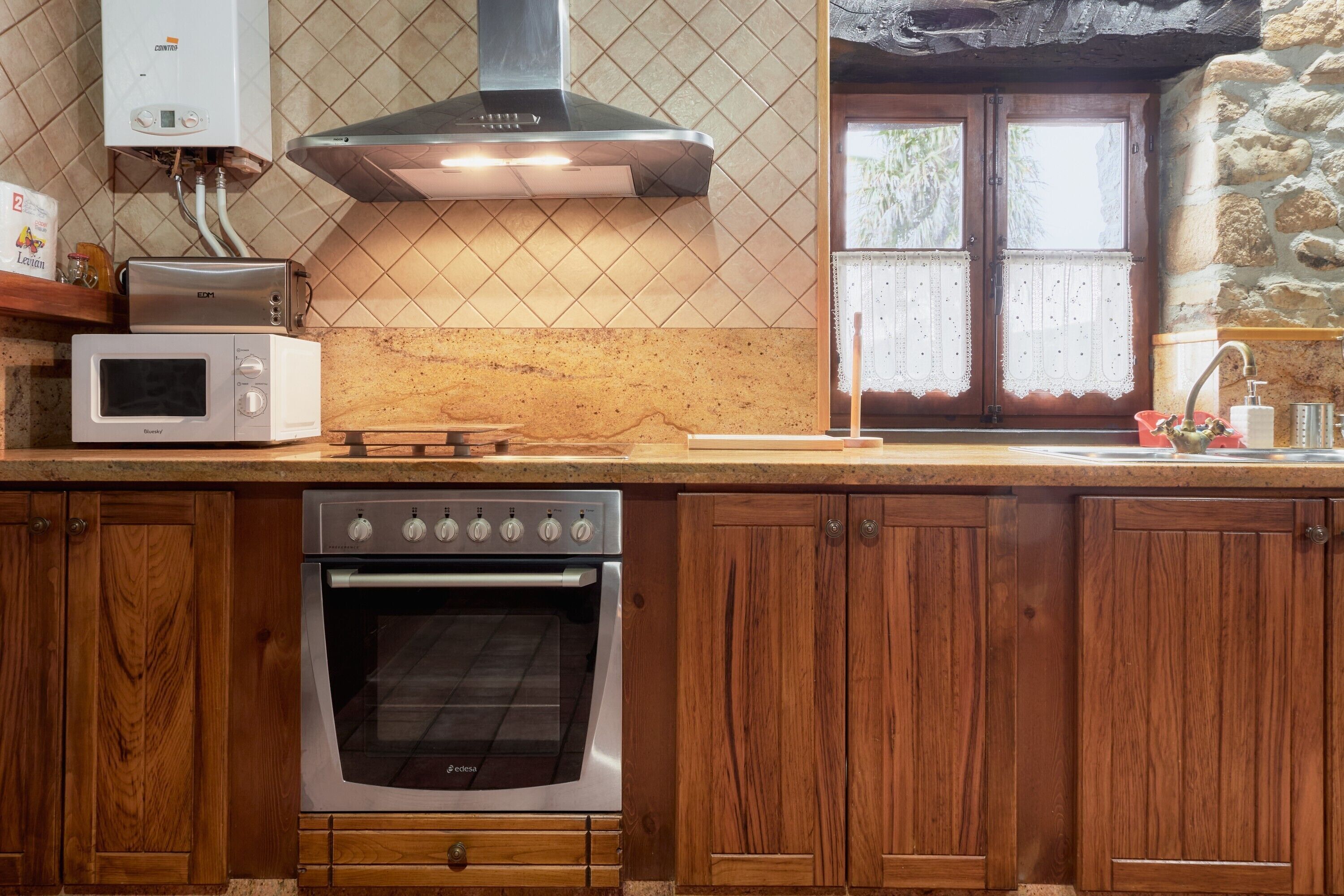 A kitchen with a built-in oven, a microwave, a gas stove, wooden cabinets, a stone wall, and a window with lace curtains.