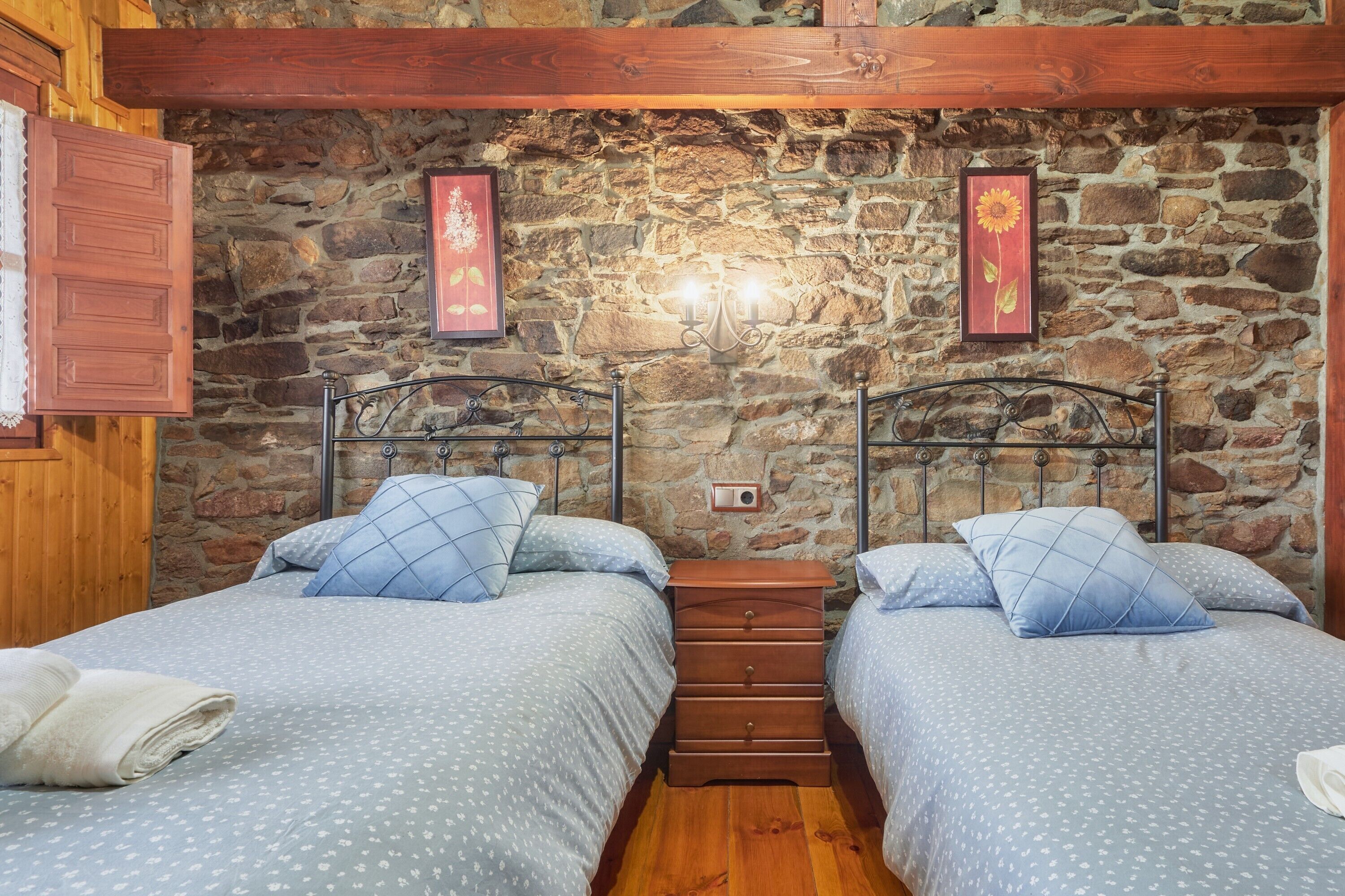 A bedroom with two beds, a stone wall, wooden beams, and a nightstand.